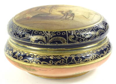 A Noritake porcelain box and cover, the lid painted with an Arabic scene of a camel, palm trees, tent etc., within gilt and cobalt blue borders, printed mark to underside, 17cm dia.