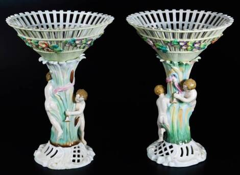 A pair of continental porcelain vase centre pieces, each with pierced dish tops, raised with flowers, on figure stems set with children, each on a pierced scroll base, polychrome decorated predominately in green and orange, unmarked, 29cm H.