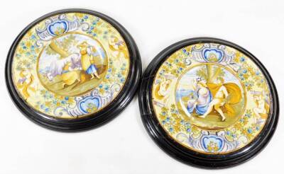 A pair of Italian Maiolica wall plaques, each of circular form, the first centred with figures and cattle, the second a classical scene with further figures with scroll borders broken by flowers and cherubs, predominately in green, orange and blue, unmar