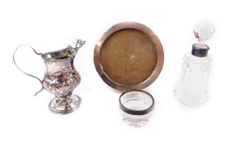 A George III silver cream jug, of baluster form, embossed with flowers and leaves, London 1784, together with a George V silver cased circular strut photograph frame, Birmingham 1911, cut glass scent bottle with silver mount, hallmarks indistinct, and a c