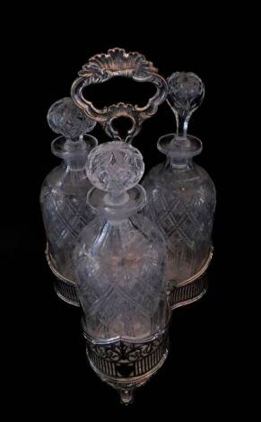 A Victorian cut glass and silver plated three bottle tantalus, the triform frame with scrolling and vertical fluted pierced decoration, raised on three scroll feet, containing three cut glass decanters, one with matched stopper, 37cm H. (AF)