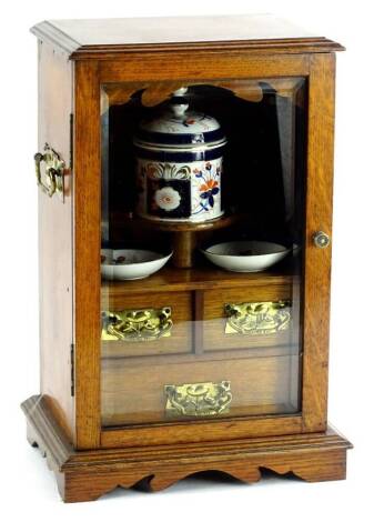 A late 19thC/early 20thC oak smoker's cabinet, the hinged bevelled glass door enclosing a porcelain tobacco jar, two dishes, each decorated in Imari colours, above two short and one long drawer, each with Art Nouveau style hammered brass handles, 43cm H, 