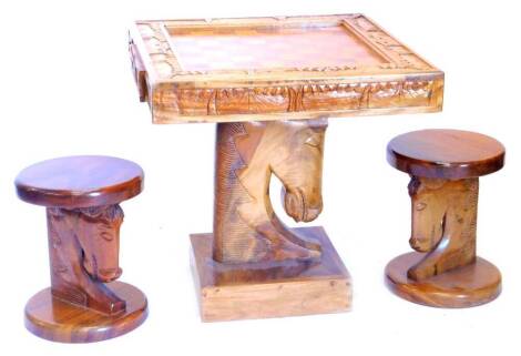 An African hardwood chess table, the square top inlaid with the board and a border carved with palm trees, buildings etc., above two frieze drawers, each containing the chess pieces, on a carved horses head support with square base, 70cm W, and two matchi