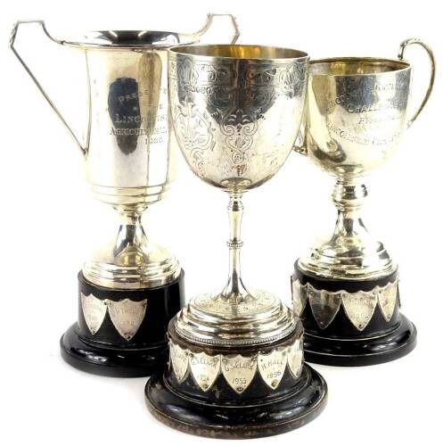 Three various silver cups, two with two handles, another goblet form with engraved decoration, each on a black base engraved with various names, 14½oz gross.