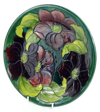 A Moorcroft pottery plate or plaque, decorated with a band of purple and pink flowers, on a green ground, impressed and hand written mark to underside, 26cm dia.