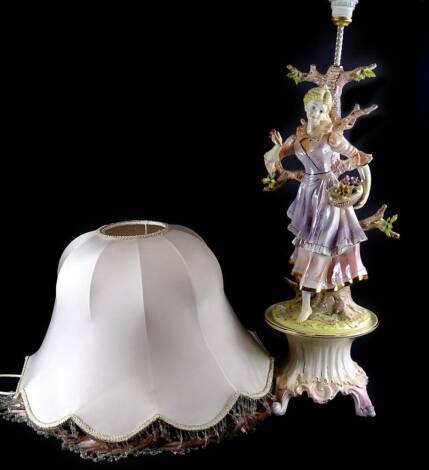 A Continental pottery table lamp, modelled in the form of a lady with basket of flowers etc., on a rococo scroll base, the shade embellished with beads etc., the lamp 85cm H.