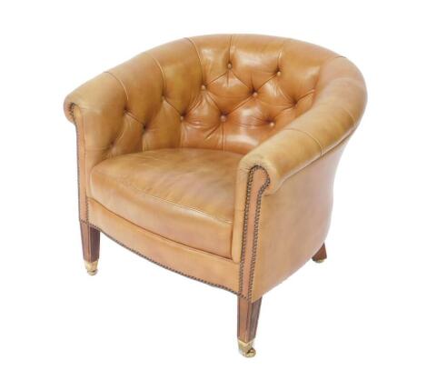 A Victorian style mahogany framed tub armchair, upholstered in button back tan leather, raised on tapering square legs brass caped on casters.