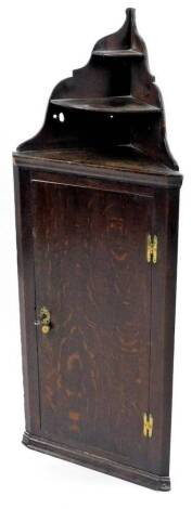 An 18thC oak corner cupboard, with a tapering raised top with two graduated shelves above a plain single plank door, moulded base with brass H hinges, 111cm H, 43cm W.