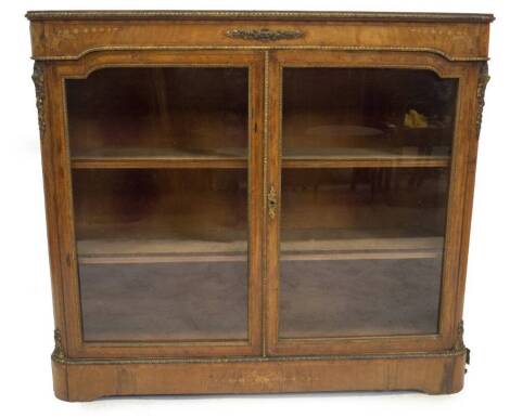 A Victorian walnut side cabinet, with rosewood and boxwood strung cross banded top, marquetry frieze with bell husk flowers and gilt metal mounts which conform to embellish the canted sides, enclosing two shaped glass doors revealing shelved interior on p