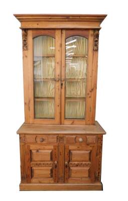 A Victorian style pine cupboard bookcase, marriage, the outswept pediment over two glazed doors enclosing three shelves, over two drawers above cupboards raised on a plinth base, 216cm H, 107cm W, 49cm D.