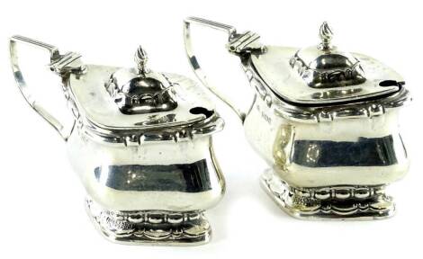 A pair of silver lozenge shaped mustard pots, each with a blue glass liner, the hinged lids with flame finials and with shaped handles, Birmingham 1908.