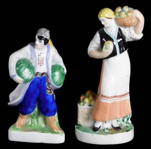 Two Russian porcelain figure groups of farmers or fruit sellers, the male figure holding melon under his arms, painted in blue, red and green, red Lomonosov porcelain mark, the other a female figure holding a basket of apples on her shoulder and another a