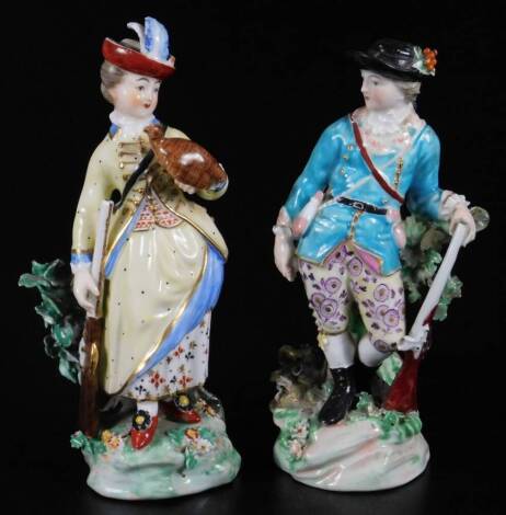 A pair of 18thC Derby Porcelain figures, of gamekeepers, each dressed in finery, he aside rifle, she holding grouse, polychrome decorated on bocage, tree bough bases, predominately in green, yellow, red and white, with gilt highlights, each with impressed