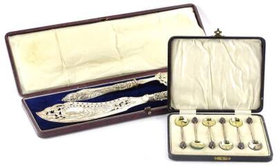 A late 19th/early 20thC silver plated fish servers, the pierced blade engraved with a fishing scene, in a fitted case, and a set of six silver gilt and green enamel coffee spoons, in a fitted case.