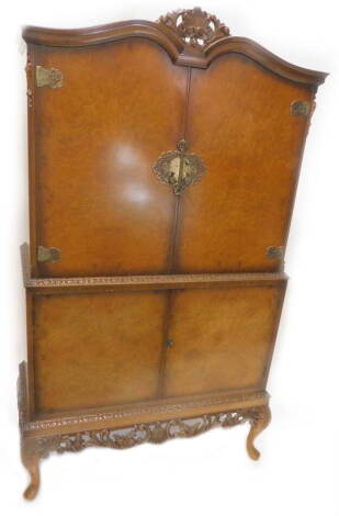 A burr walnut cocktail cabinet, the top with a shaped crest, above two doors, each with elaborate brass hinges, enclosing a glass and mirrored interior, above two further doors, enclosing shelves, with a pierced and carved apron on cabriole legs, 97cm W.