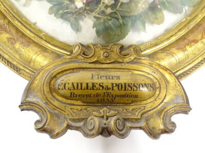 A mid 19thC French silk flower display, representing roses, leaves and other similar flowers, in oval gesso frame, the crest decorated with a bow, etc., exhibition plaque titled Fleurs, ACAILLES de POISSONS, brevet de l'exposition 1855, 61cm x 41cm. - 2