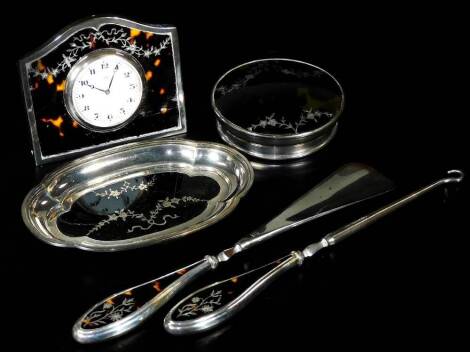 A George V silver and tortoiseshell part dressing table set, comprising of a travel clock, with 4.5cm Dia. Arabic dial, marked 8 days, with easel back, a shaped tray, button hook, shoe horn and lid, Birmingham 1925. (a quantity)