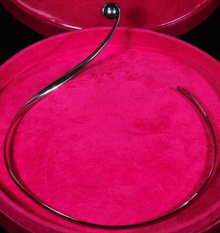 A Paul Spurgeon 18ct white gold and cultured pearl necklace, designed as a ribbon of gold curled around a dark bluey/grey cultured pearl, tension set to turn freely, London assay, in designer's fuschia leather circular box, approx 42.8g all in.