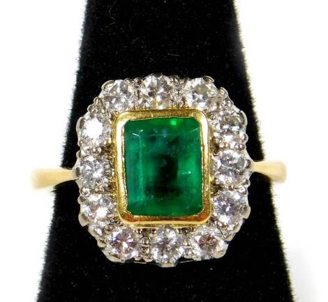 An 18ct gold emerald and diamond dress ring, with central rectangular cut emerald 6.8mm x 5.4mm x 4.6mm, approx 1.19cts, in rub over setting, surrounded by twelve round brilliant cut diamonds, each in platinum claw setting, maker D.P, Birmingham assay, ri