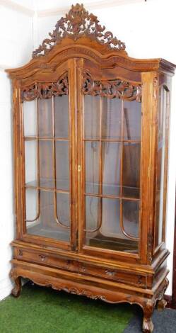 A continental walnut display cabinet, the pierced scroll and floral finial raised above two astragal glazed doors revealing shelves with two drawers beneath, on compressed cabriole legs joined by a carved lower section, 117cm H, 118cm W, 40cm D.