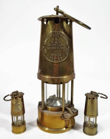 An Eccles Protector brass miner's lamp, of cylindrical form with glass centre and hook top, 26cm H, and two further miniature miner's lamps. (3)