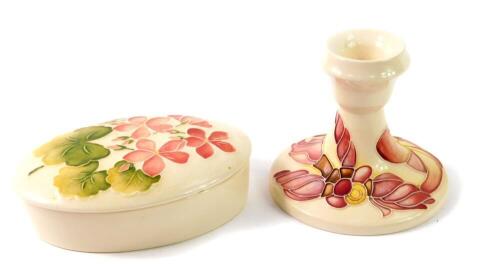 A Moorcroft pottery oval box and cover, decorated in the Pink Geranium pattern, impressed marks, 13cm W, together with a candlestick of squat form decorated in the Magnolia pattern, impressed and painted marks, 9cm H. (2)