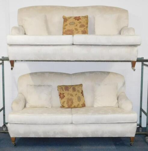 A pair of Edwardian style two seater sofas, upholstered in a floral pattern cream fabric, raised on tapering square legs, brass capped on castors, 189cm W.