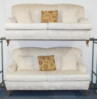 A pair of Edwardian style two seater sofas, upholstered in a floral pattern cream fabric, raised on tapering square legs, brass capped on castors, 189cm W.
