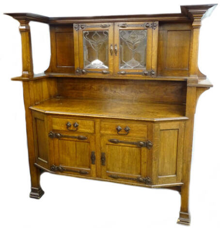 A late 19thC oak Arts and Crafts sideboard in the manner of Liberty, the raised back with two leaded glazed doors, each with hammered copper hinges and fittings, the bow fronted base with two drawers above two doors, with similar hammered fittings, on blo