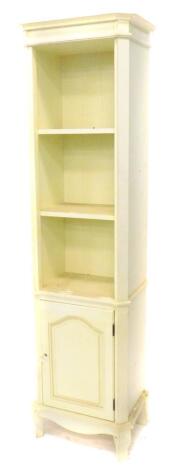 A narrow cream painted display unit, with a moulded cornice above three shelves and a panel door on scroll feet, 187cm H, 50cm W.