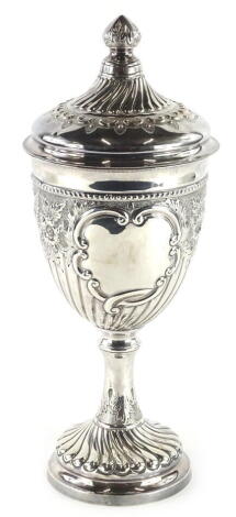 A late Victorian silver trophy cup and cover, the lid with a turned finial and with fluted decoration with embossed and repousse 'C' scrolls, within a beaded border, the base heavily embossed with flowers, fruits etc., surrounding two vacant cartouches, 