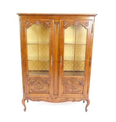 An 18thC style oak display cabinet, carved with rococo scrolls and leaves, having two glazed doors opening to reveal two glass shelves, raised on cabriole legs, 158cm H, 115cm W, 37cm D.