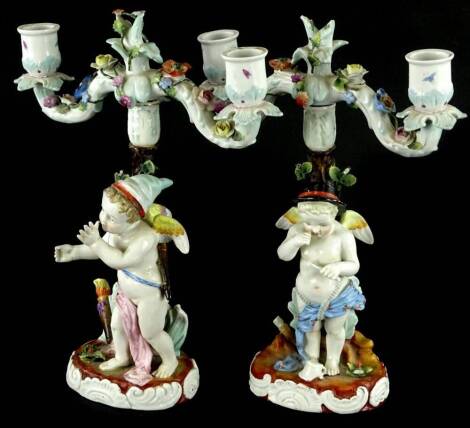 A pair of late 19thC German porcelain two handled candelabra, each modelled with a putto wearing a turquoise and a black hat, the flower encrusted branches each with two sconces, on a domed foot, blue mark to underside, 29cm H.