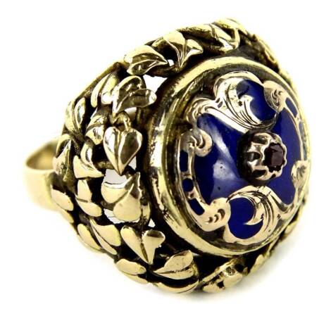 An 18ct gold enamel designed dress ring, with central circular design centre with ruby set stone, and scroll border design, with leaf moulded sides and borders, stamped 750, ring size M, 7.5g all in.