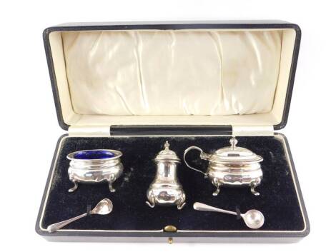 A George V silver three piece condiment set, of fluted form, raised on paw feet, with blue glass liners, comprising salt, mustard pot and pepperette, together with a salt spoon, cased, Adie Brothers Ltd, Birmingham 1931/32, 4.61oz.