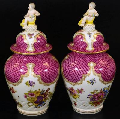 A pair of Meissen porcelain vases and covers, each exquisitely decorated with flowers, with domed circular lids surmounted by cherub and garland finials, each main body decorated with feathered pink and purple panelling, with further handpainted flowers o