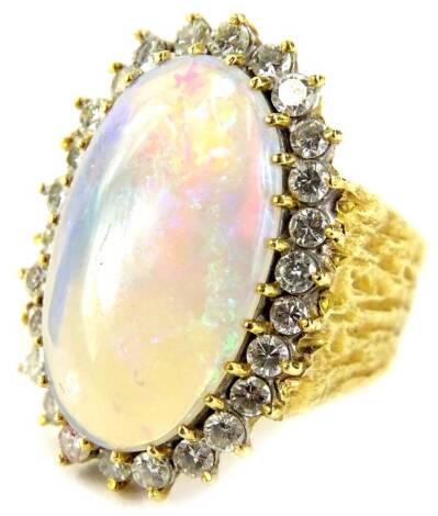 An opal and diamond dress ring, set with central elongated oval imitation opal 2cm x 1.5cm, surrounded by twenty six diamonds, over two tiers, on a hammered yellow metal band, with faint rubbed markings, possibly 18ct, ring size Q, 14.2g all in.