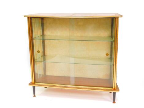 A vintage 1950's teak display cabinet, of trapezium form, with two glazed doors opening to reveal two glass shelves and decorated glass side panels, floral fabric back, the front with brass and ebonised trim, raised on turned ebonised legs on brass caps, 