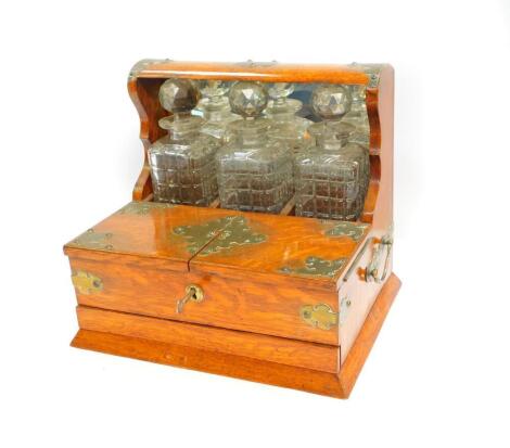 An Edwardian oak and brass bound three bottle tantalus and cigar box, the mirrored back tantalus containing three cut glass decanters and stoppers, fronted by a box with two hinged lids opening to reveal a fitted compartment, above a frieze drawer, on a 