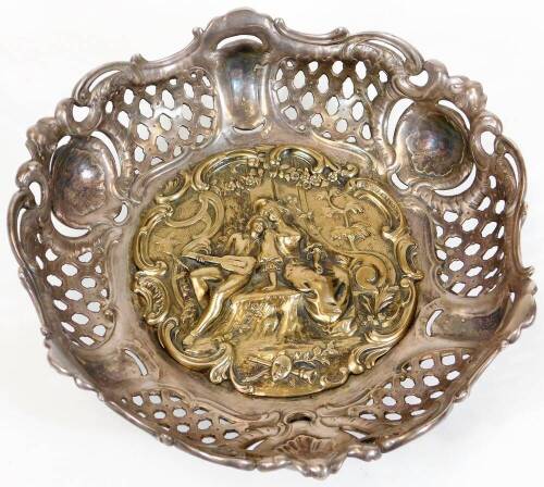 A late 19thC continental white and gilt metal bowl, the central plaque embossed with courting figures within floral garlands and rococo scrolls, within a rococo scroll and lattice border, raised on three leaf scroll feet, 26cm diameter.