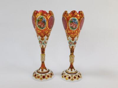 A pair of late 19thC Bohemian flashed cranberry glass vases, of ogee form, raised on a slender collared stem, reserve painted with flowers, against a white ground, with gilt foliate decoration against the cranberry glass, 26.5cm H. (AF)