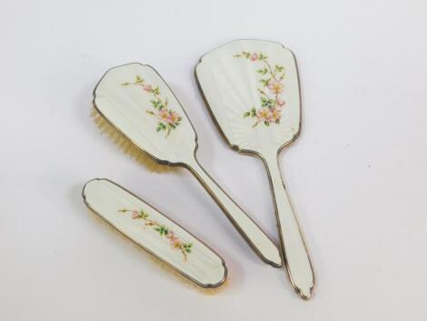 A silver and enamel hairbrush, hand mirror and clothes brush, guilloche decorated, painted with flowers against a white enamel ground, Mappin & Webb, Birmingham 1964. (3)