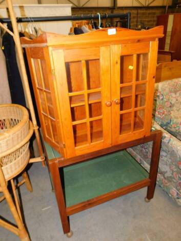 A pine and glazed wall mounted cabinet, 75cm wide, together with a teak trolley with two removable green plastic tray inset, 76cm wide. (2)