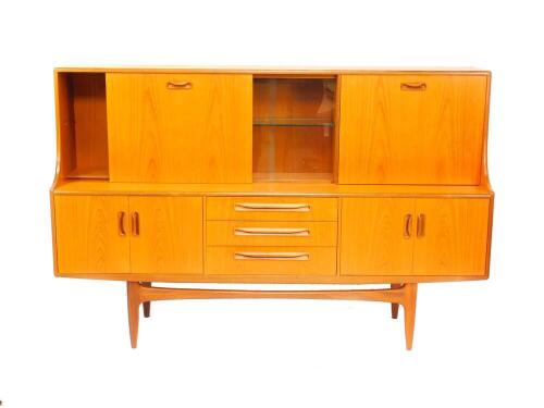 A G-Plan Fresco teak sideboard, with a pair of glazed central doors opening to reveal a lit single shelf unit, flanked by a drop down flap drinks cabinet, and sliding door opening to reveal a single shelf compartment over three drawers flanked by two pai