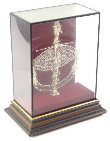 A white coloured metal filigree and wirework centre piece, of elaborate ellipse form on a circular foot, in Perspex case on wooden stand, the case 33cm high.