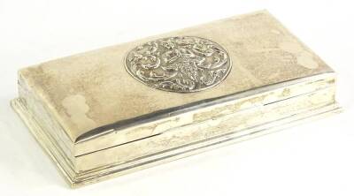 A white metal cigarette case, of rectangular plain form, the thumb moulded handle of plain form, raised with a panel of a god and scrolls to the centre, with fitted interior, on a stepped base, marked Sterling, 22cm wide, 14oz all in.