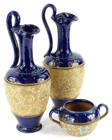 A pair of Royal Doulton Slater's patent ewers, each decorated with a central band of flowers and leaves embellished with coloured enamels on a gilt ground, with cobalt blue bands to top and bottom, 32cm high, and a two handled sugar bowl.