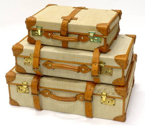 A graduated set of three canvas and brown leather vintage suitcases, the largest 73cm wide, smallest 47cm wide.