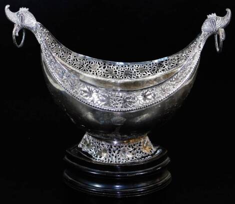 An early 20thC Indian ceremonial bowl, of shaped ellipse form with heavily cast ring handles, and a pierced border set with flowers, on an oval foot, filigree decorated with further flowers heads, on a removable wooden plinth base, 31cm high, 38cm wide.