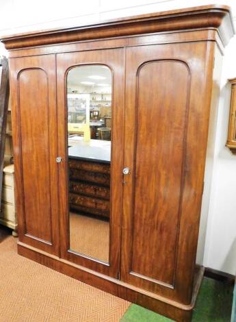 A Victorian mahogany triple door wardrobe, with moulded cornice, raised above a central mirror flanked by two panelled doors on a plain block base, 210cm high, 200cm wide, 59cm deep.